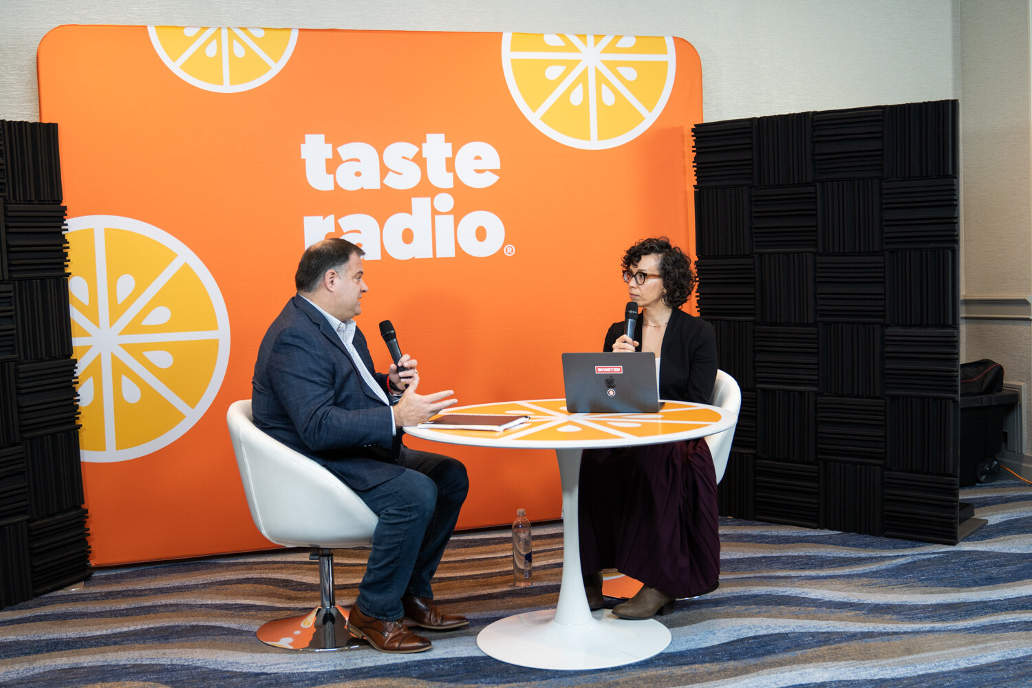 Two people talk at a small table in front of a Taste Radio backdrop.