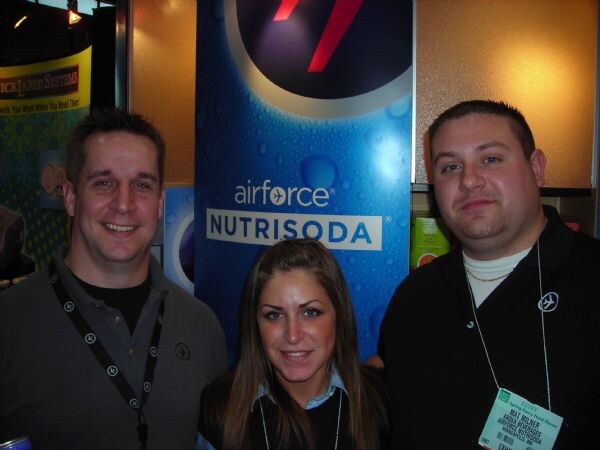 Two men and a woman pose together in front of a blue Airforce Nutrisoda banner. Both men wear lanyards and the man at right has a visible exhibitor badge.