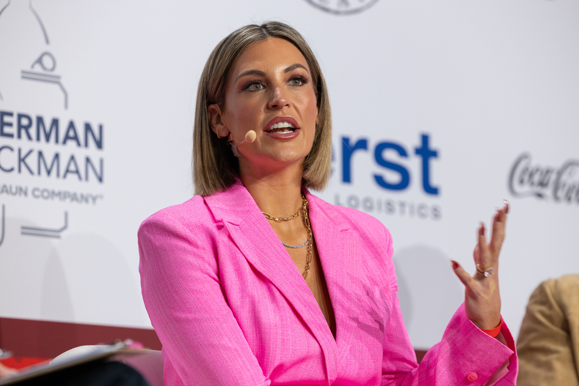 A woman in a pink blazer gestures while speaking on stage.