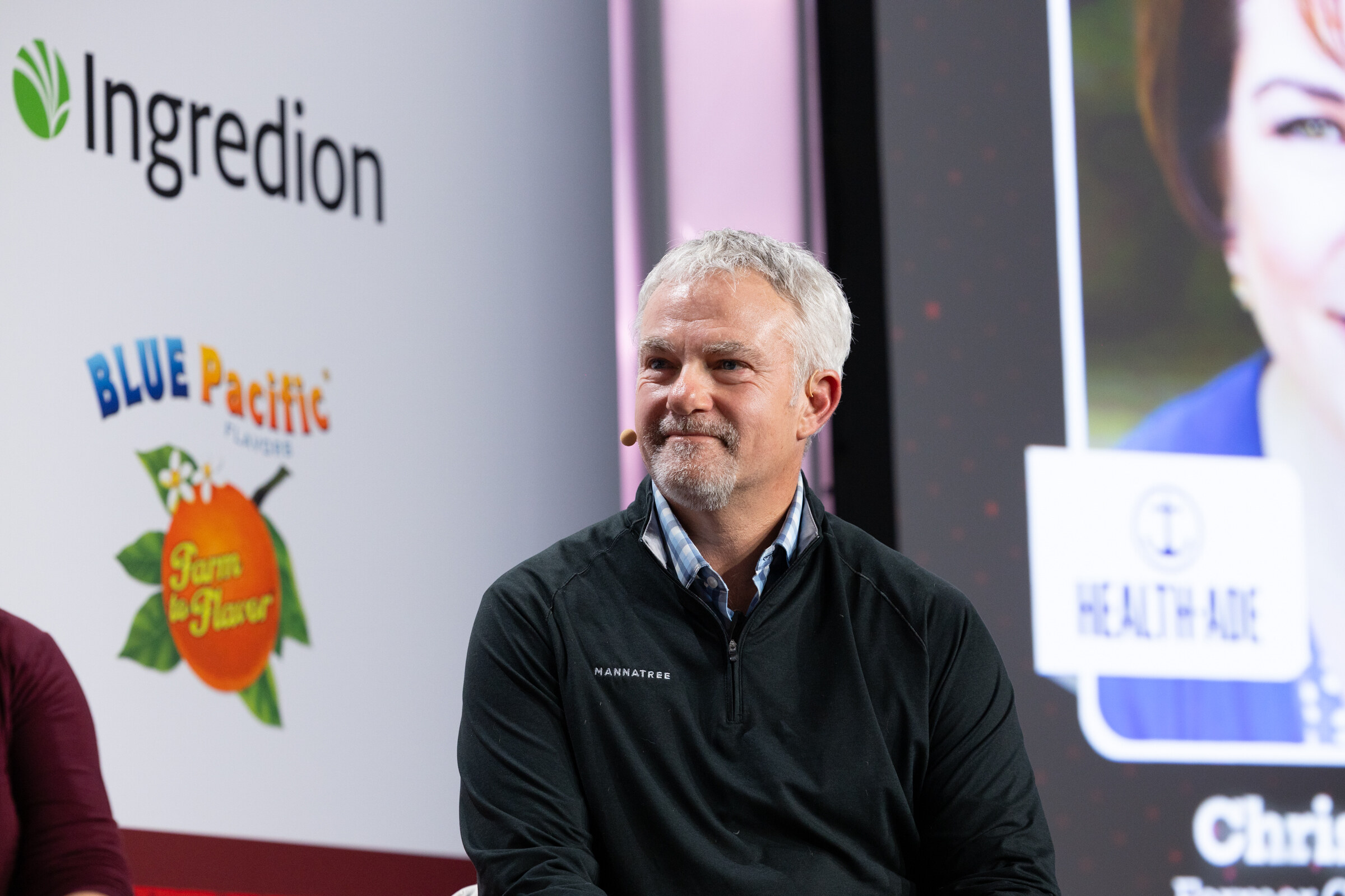 A man in a black quarter-zip sits on stage in front of a Health-Ade slide.