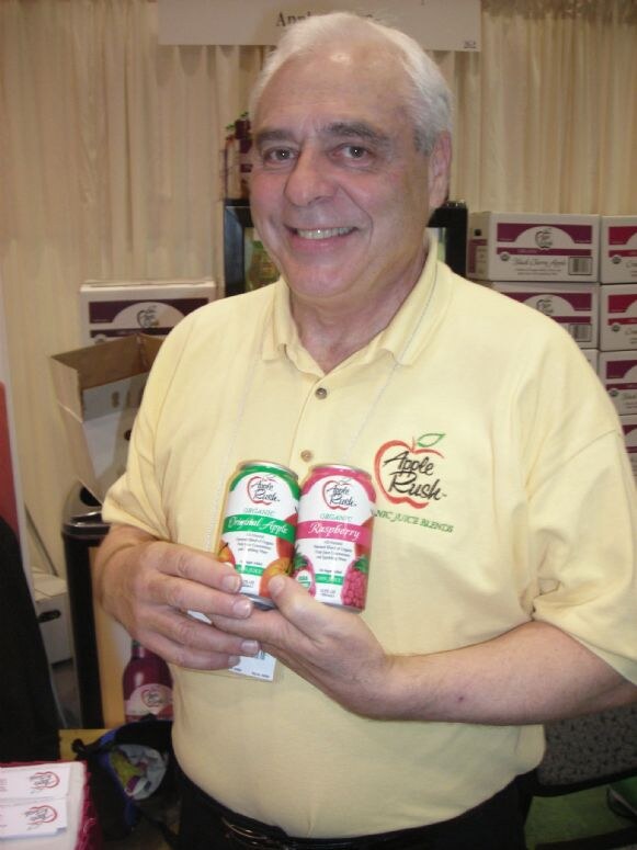 A smiling man in a yellow Apple Rush polo holds two cans of organic juice, Original Apple and Raspberry, with boxes of juice stacked behind him.