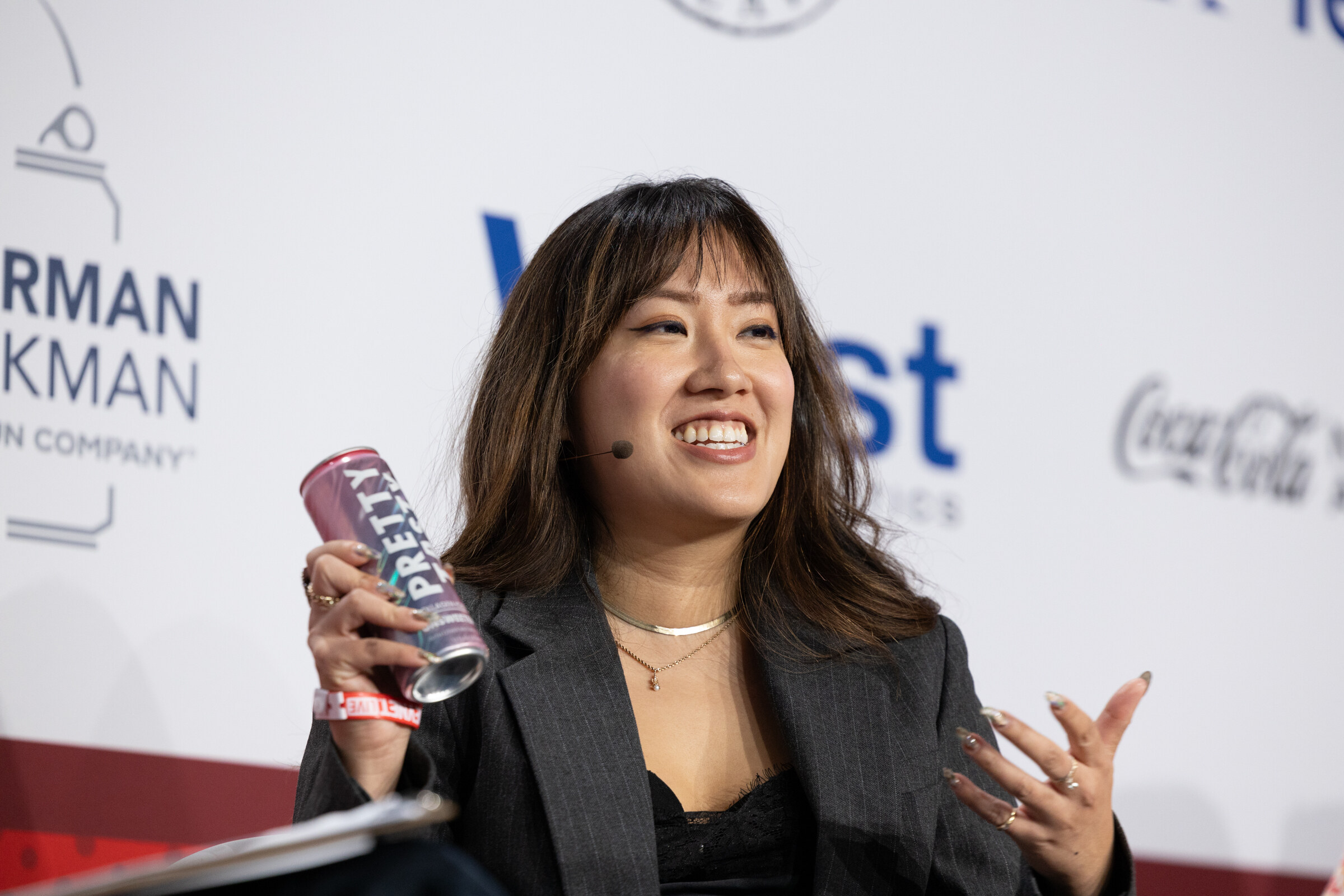 A woman in a gray blazer holds up a can while speaking.