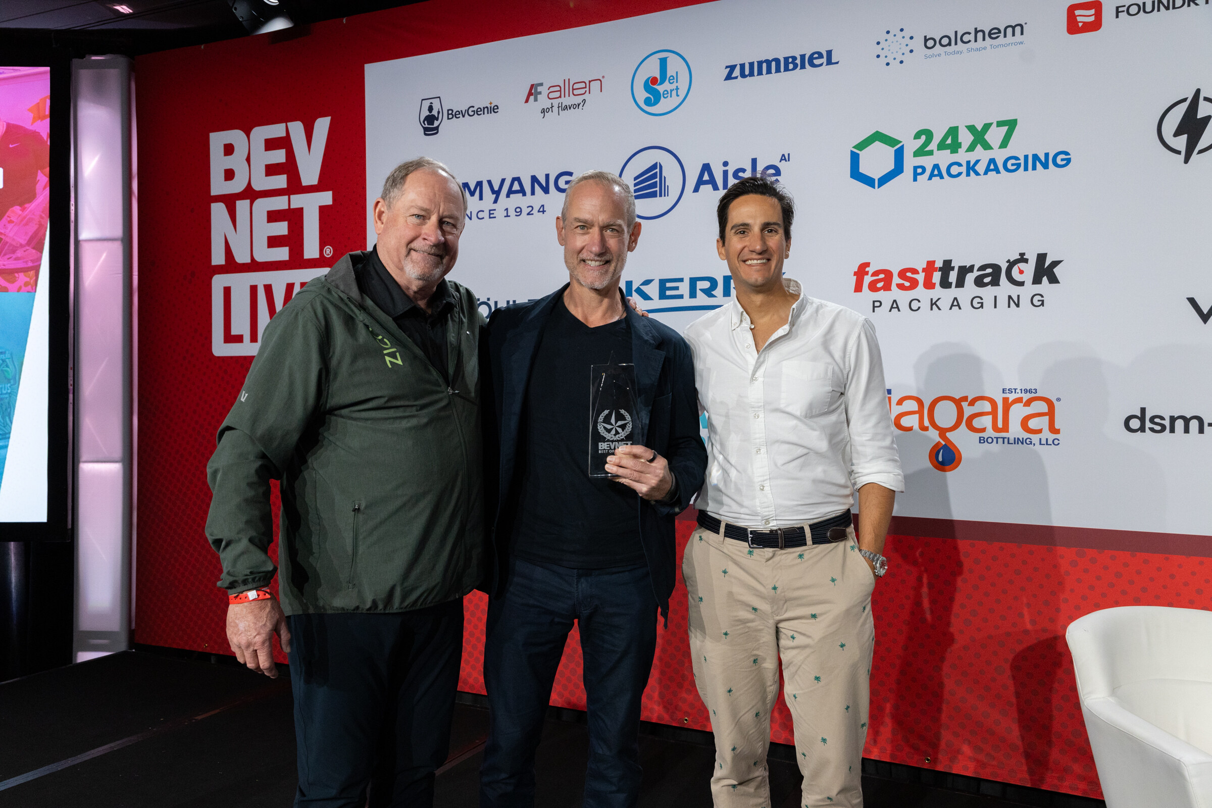 Three men pose on stage, one holding a glass award.