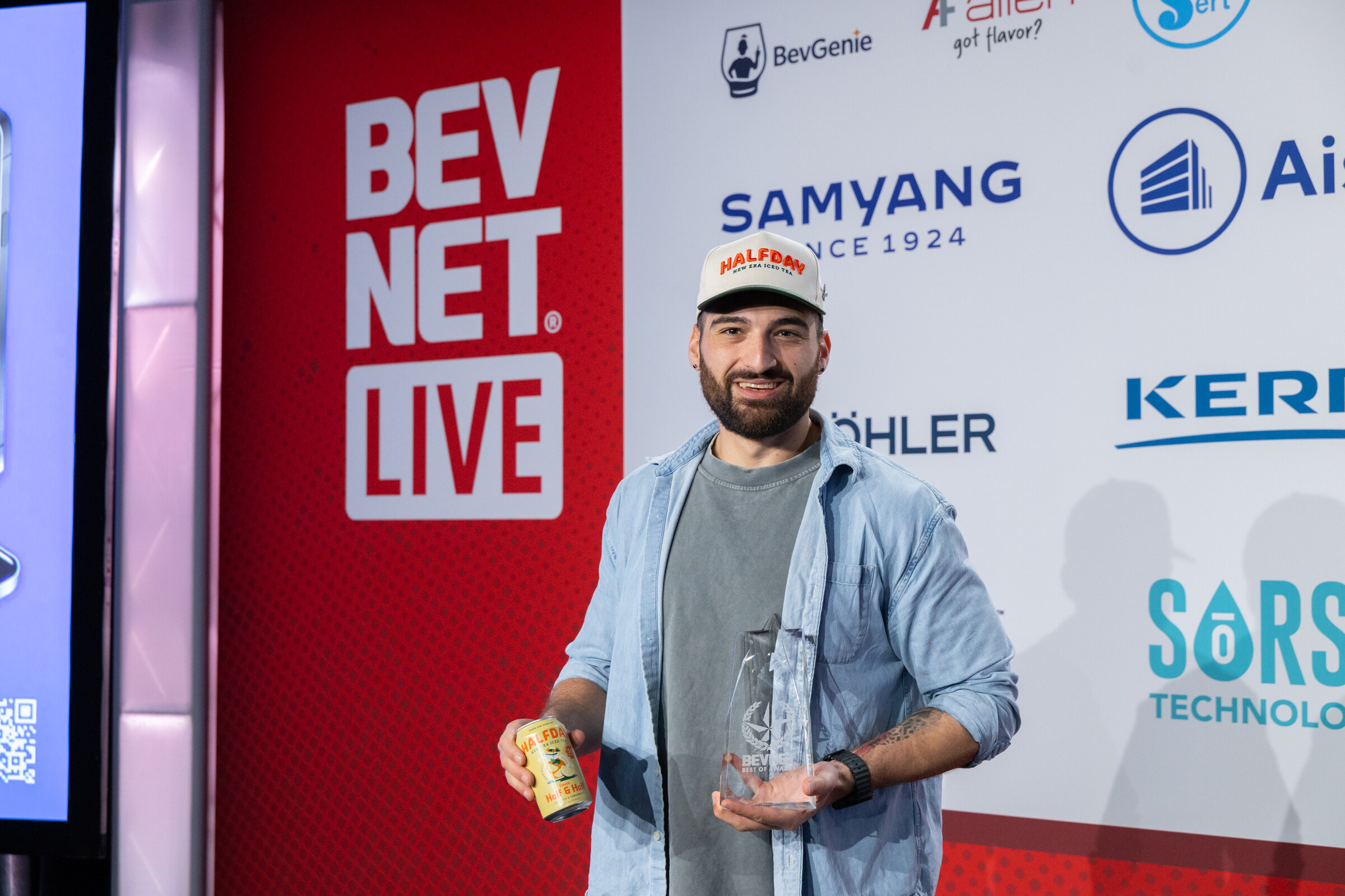 A man in a cap holds a glass award and a can on stage.