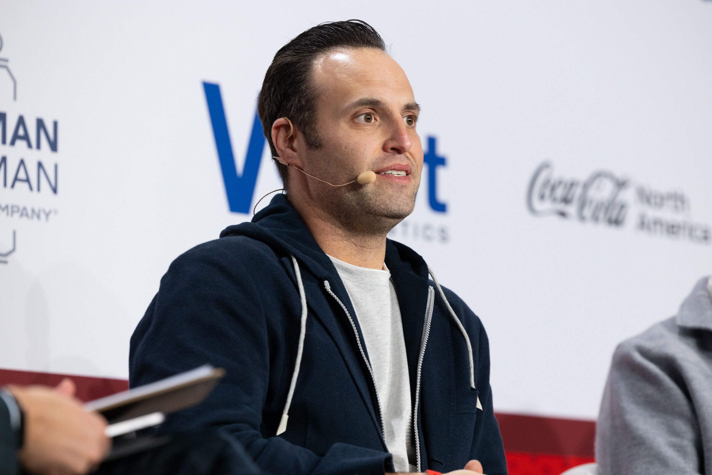 A man in a navy hoodie speaks into a headset microphone on stage.