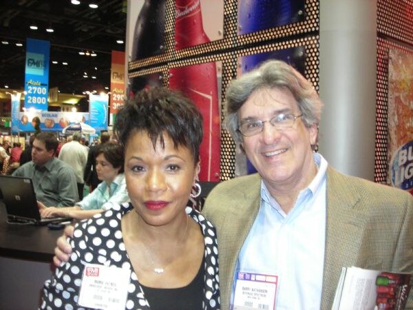 Barry Nathanson of Beverage Spectrum, wearing glasses and a tan blazer, stands beside a woman in a polka-dot jacket in front of Budweiser bottle graphics.