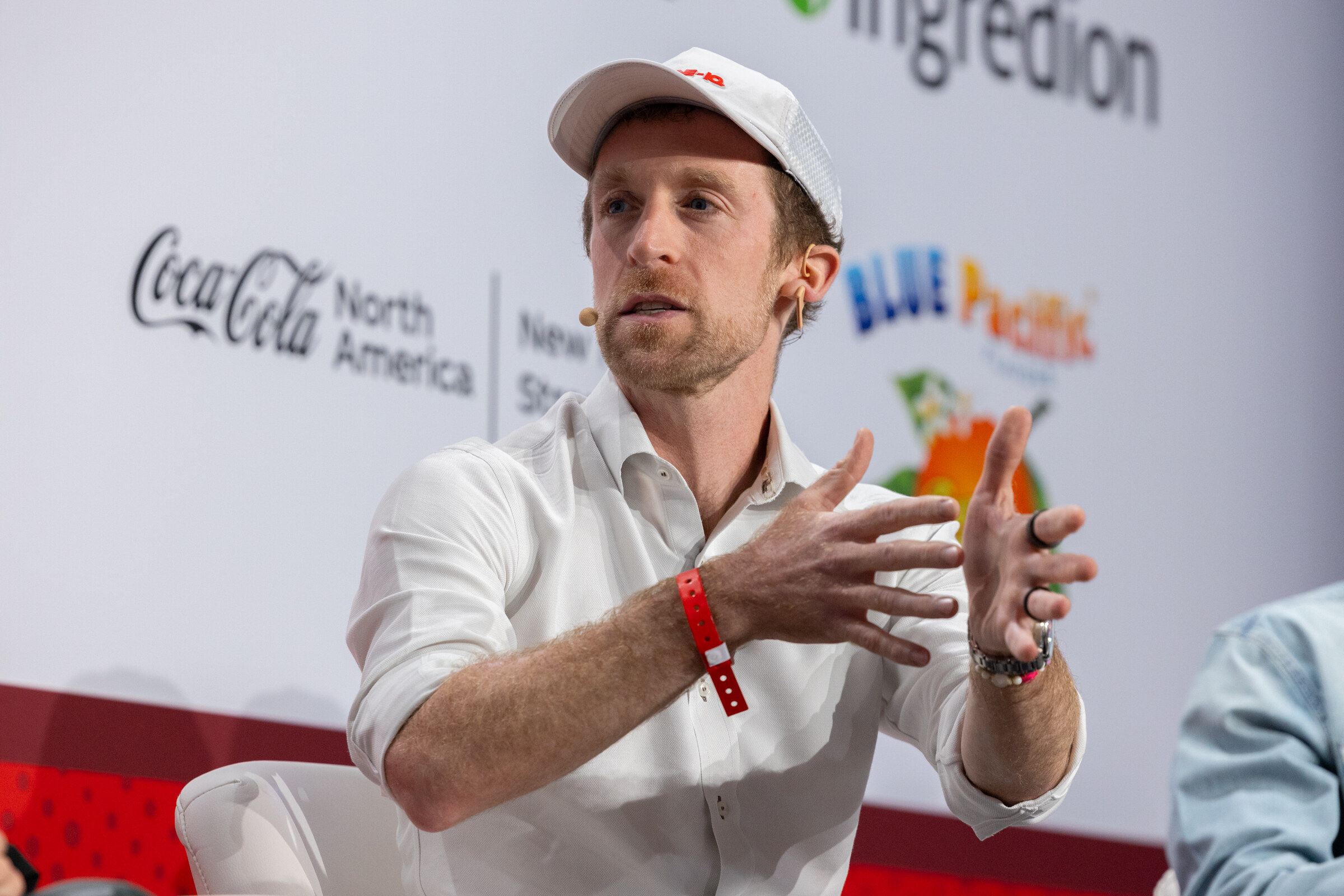 A man in a white shirt with a cap on the table gestures while talking.