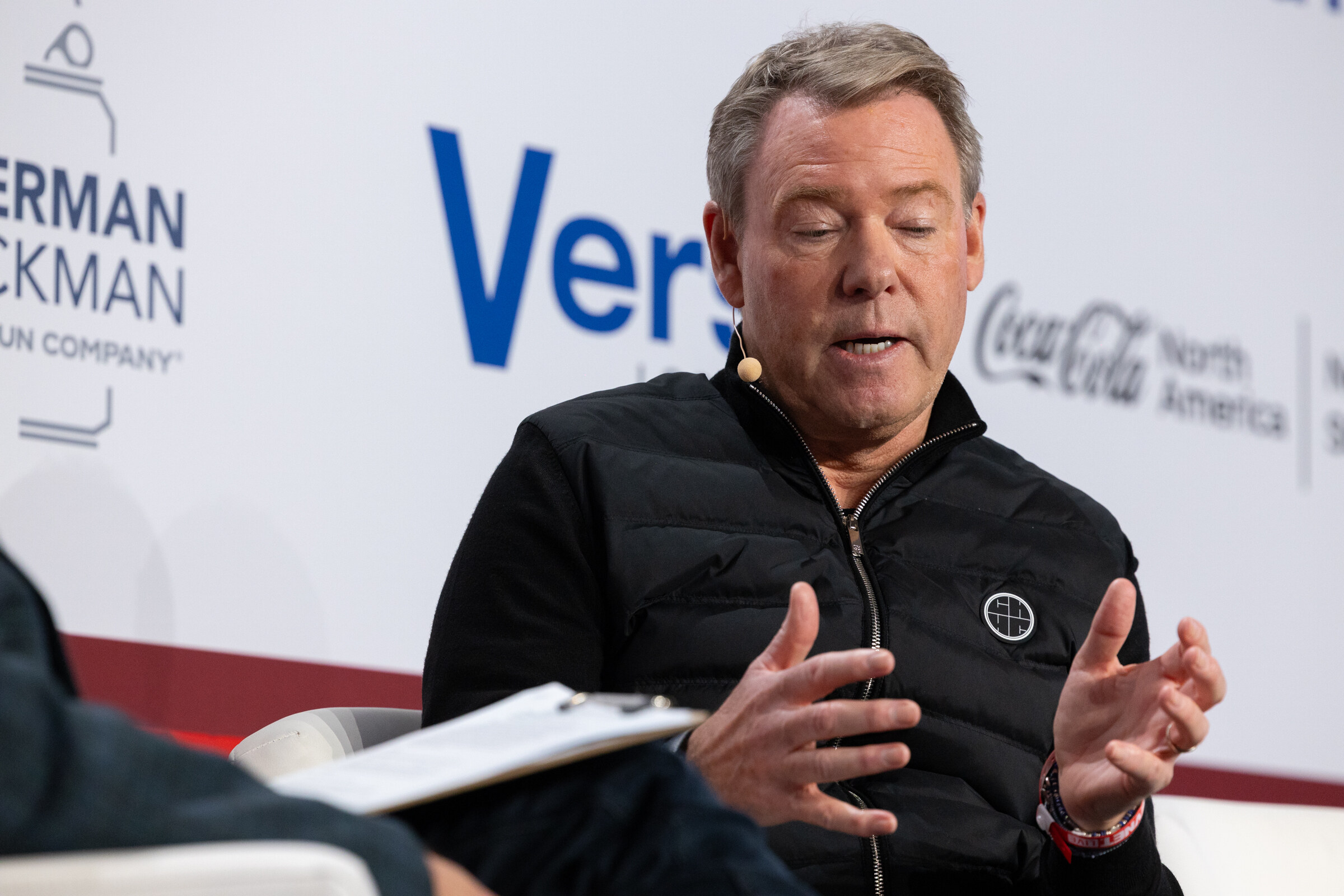 A man in a black quilted vest speaks with a clipboard on his lap.