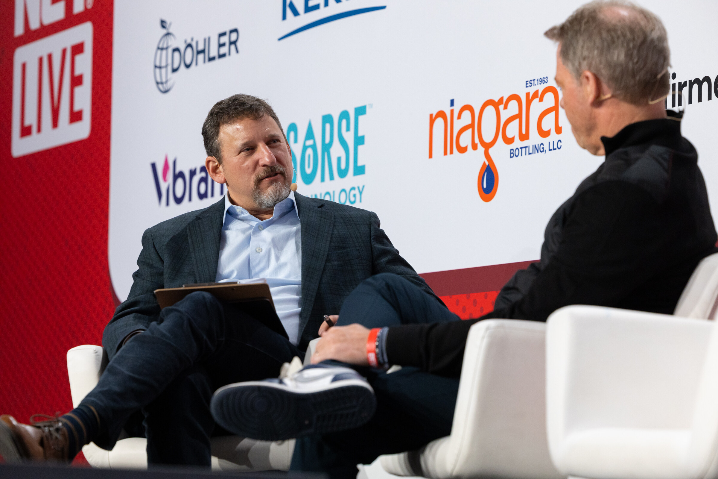 Two men sit in white chairs on stage, one holding a notepad.