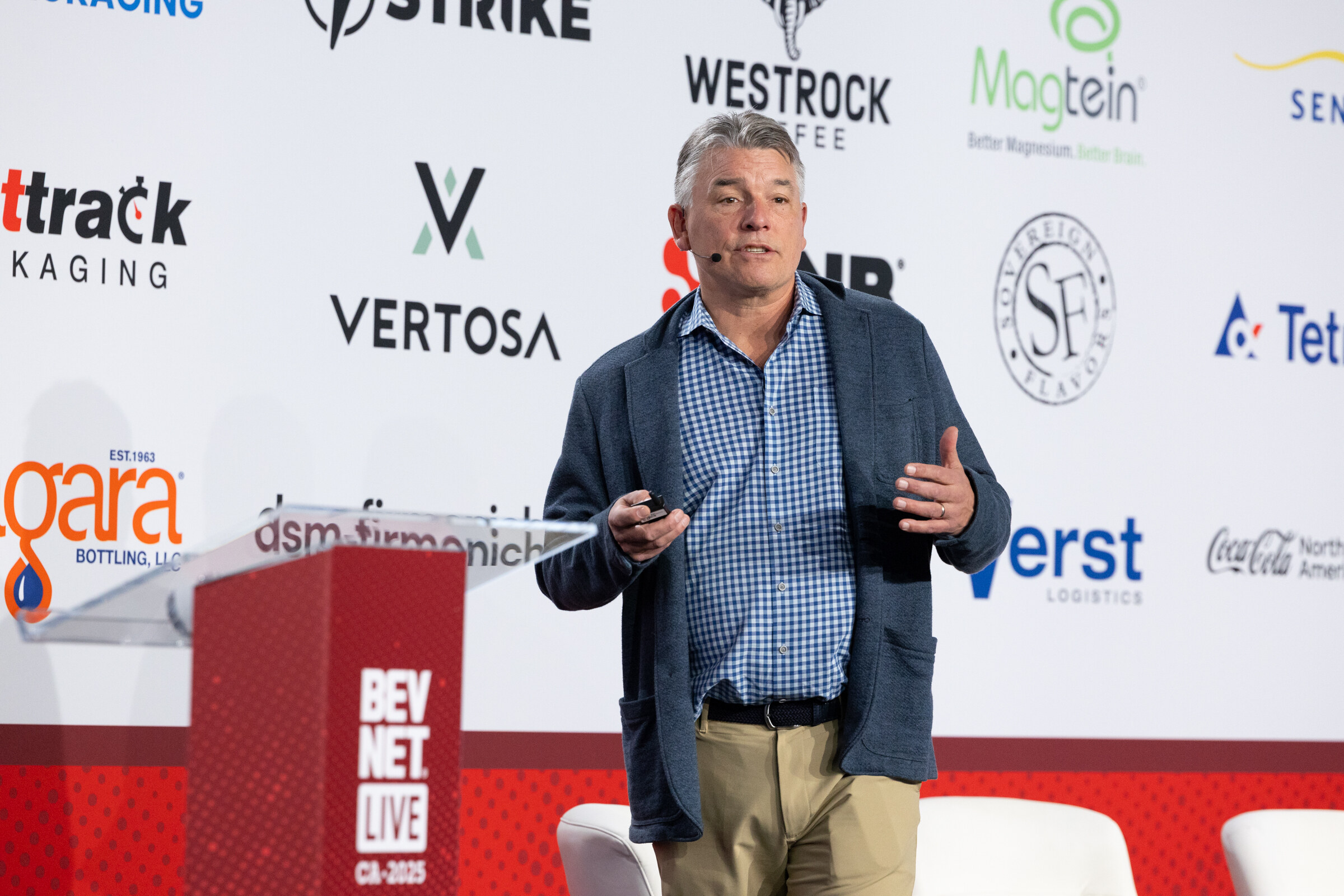 A man in a blue jacket speaks on stage in front of a sponsor wall.