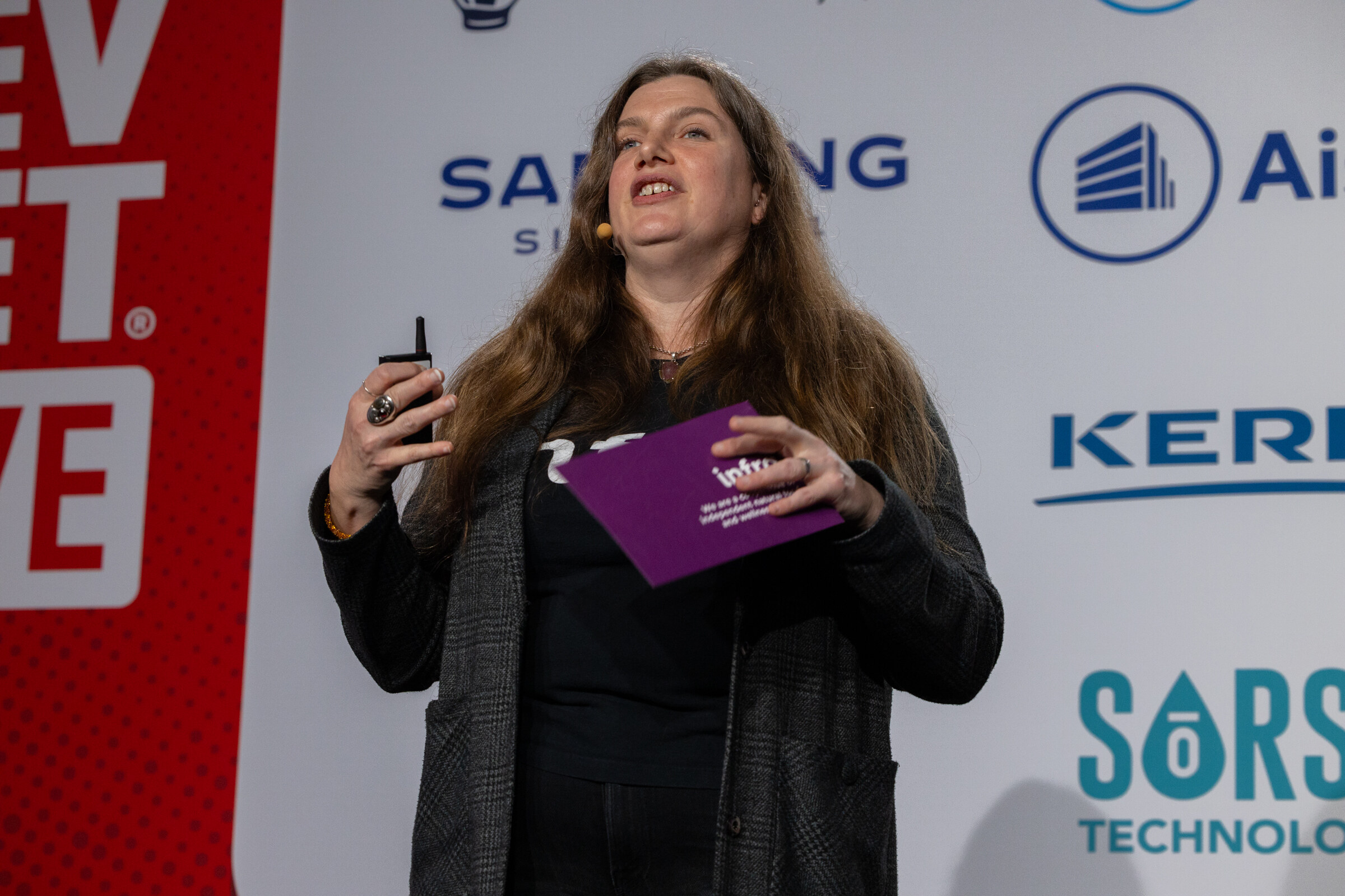 A woman in a black INFRA shirt holds notes and speaks on stage.