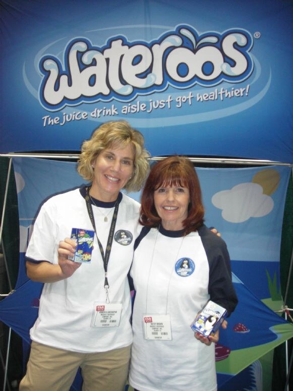 Betsy Brown, right, and another woman, left, in matching white Wateroos shirts each hold a juice drink box in front of a blue Wateroos banner.