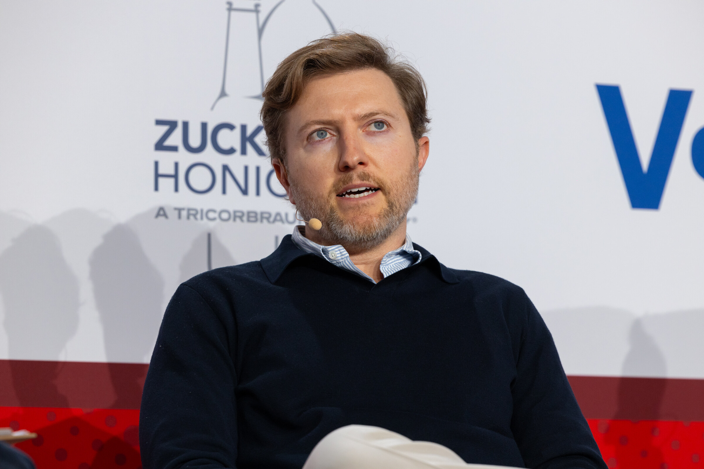 A man in a navy sweater listens while seated on stage.