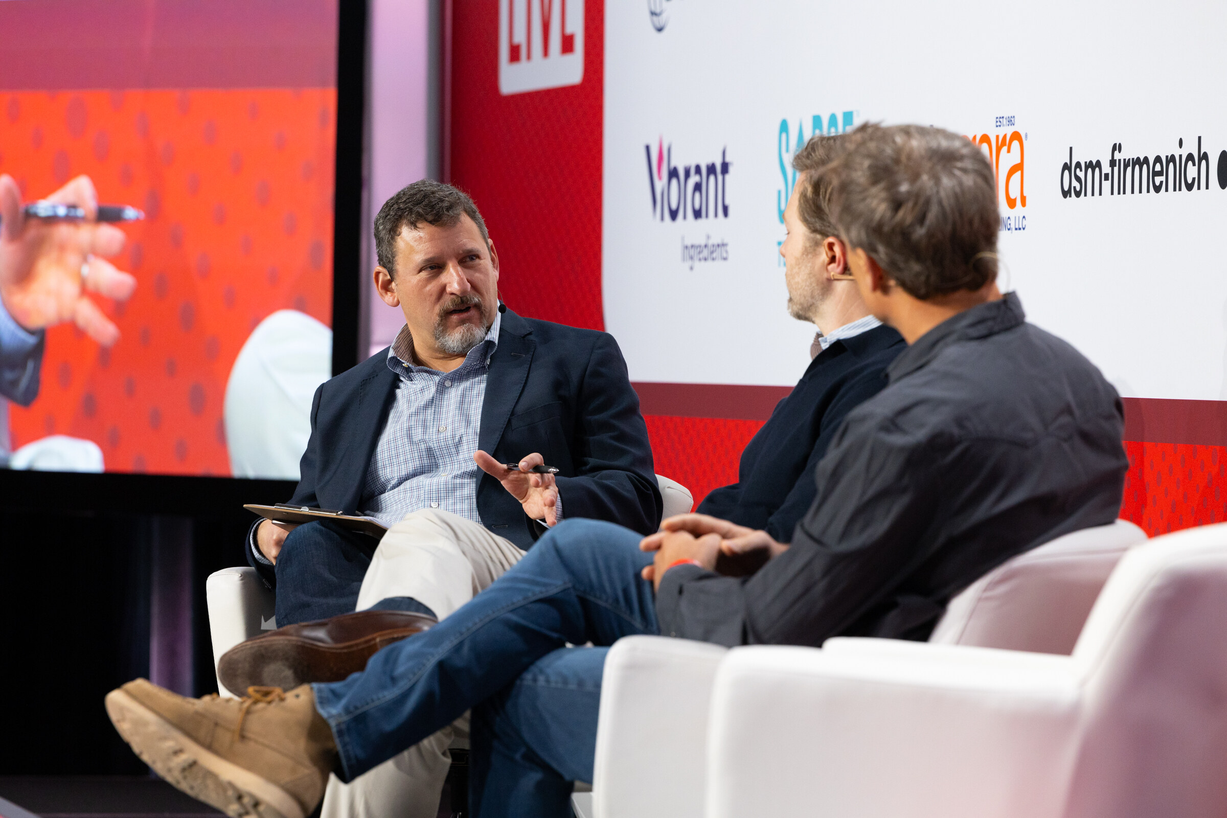 Three men sit in white chairs on stage in conversation.