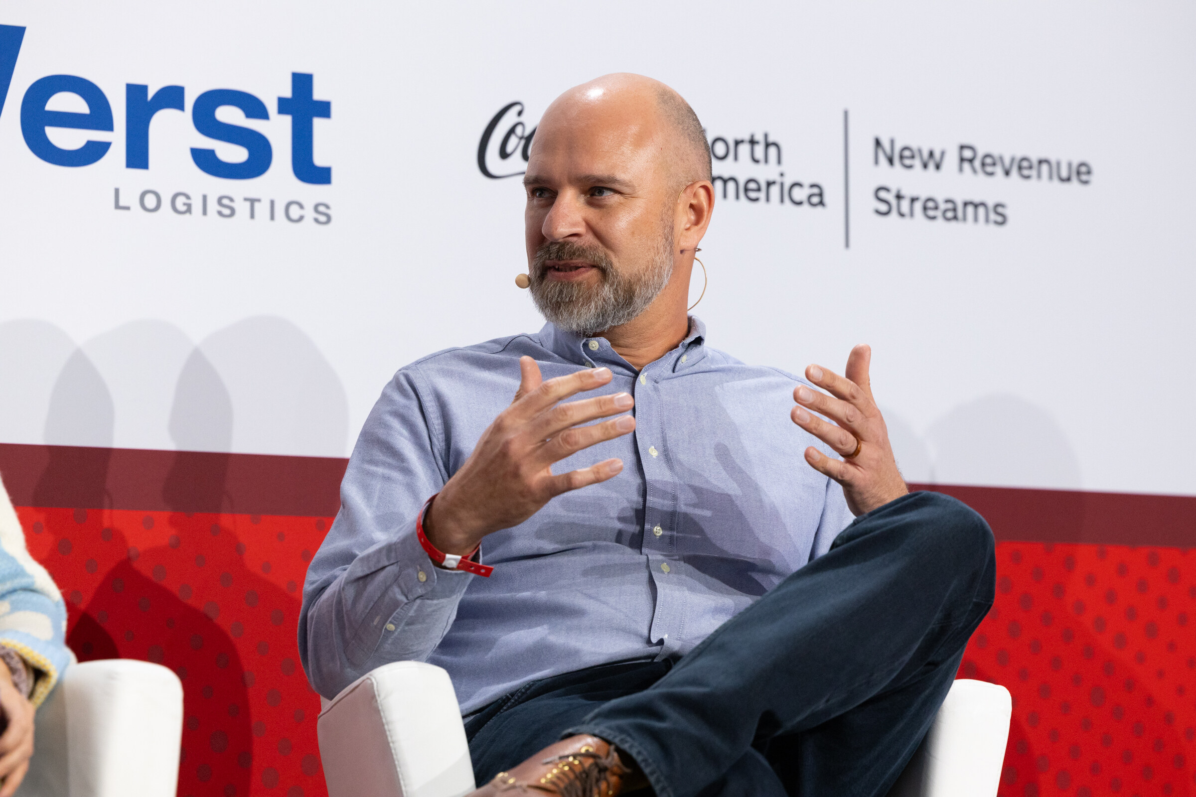 A bald man in a blue shirt gestures while seated.