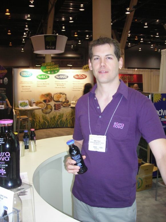 A man in a purple Bossa Nova polo holds a small bottle of juice at a curved counter displaying more Bossa Nova bottles, with other exhibitor booths behind him.
