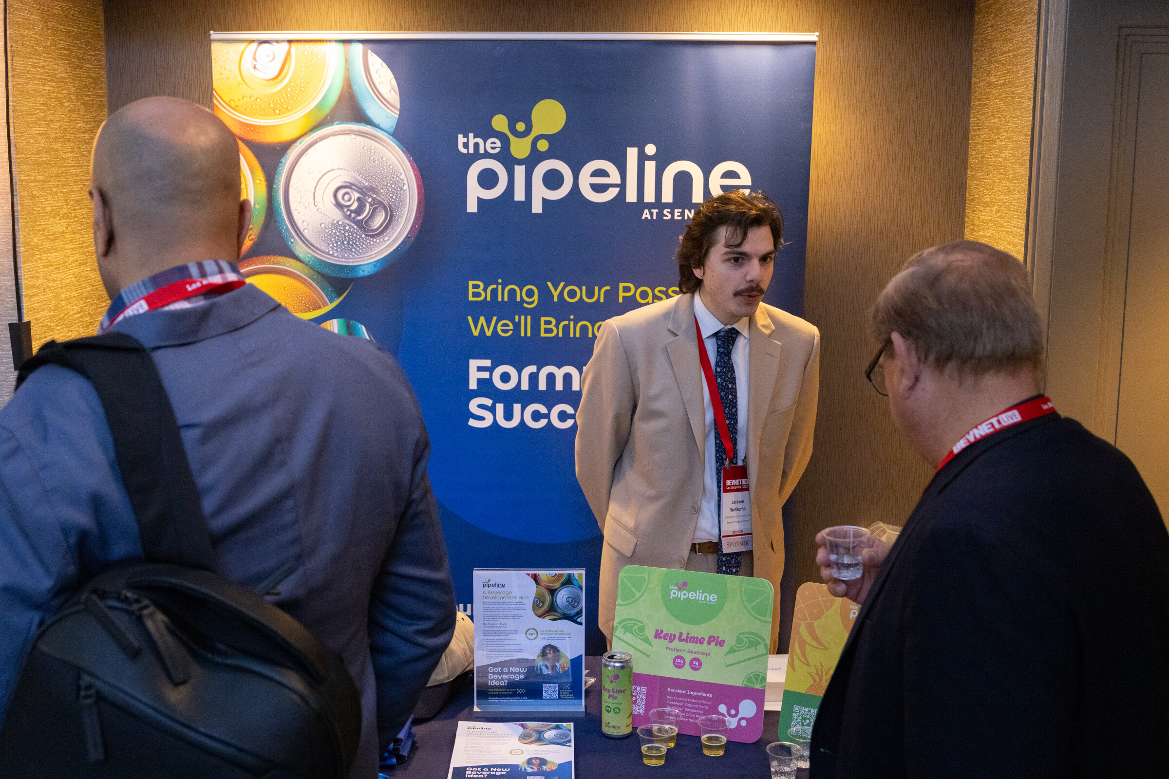 A man in a tan suit stands at a Pipeline booth with visitors.