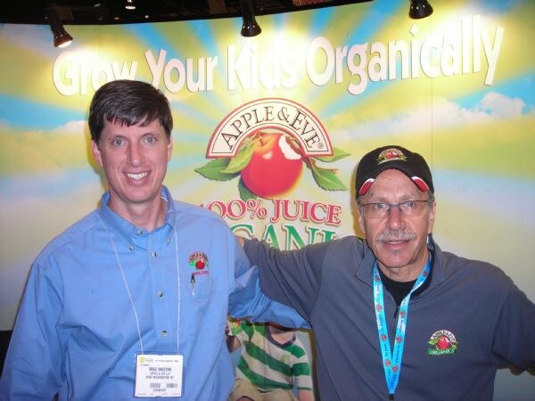 Brad Angevine of Apple & Eve LLC, in a blue shirt, stands beside a man in a black Apple & Eve cap in front of a booth backdrop reading Grow Your Kids Organically.
