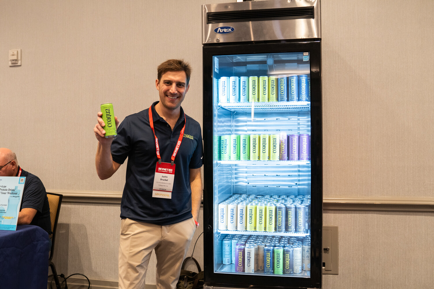 A man in a navy polo holds up a green can beside a lit fridge.