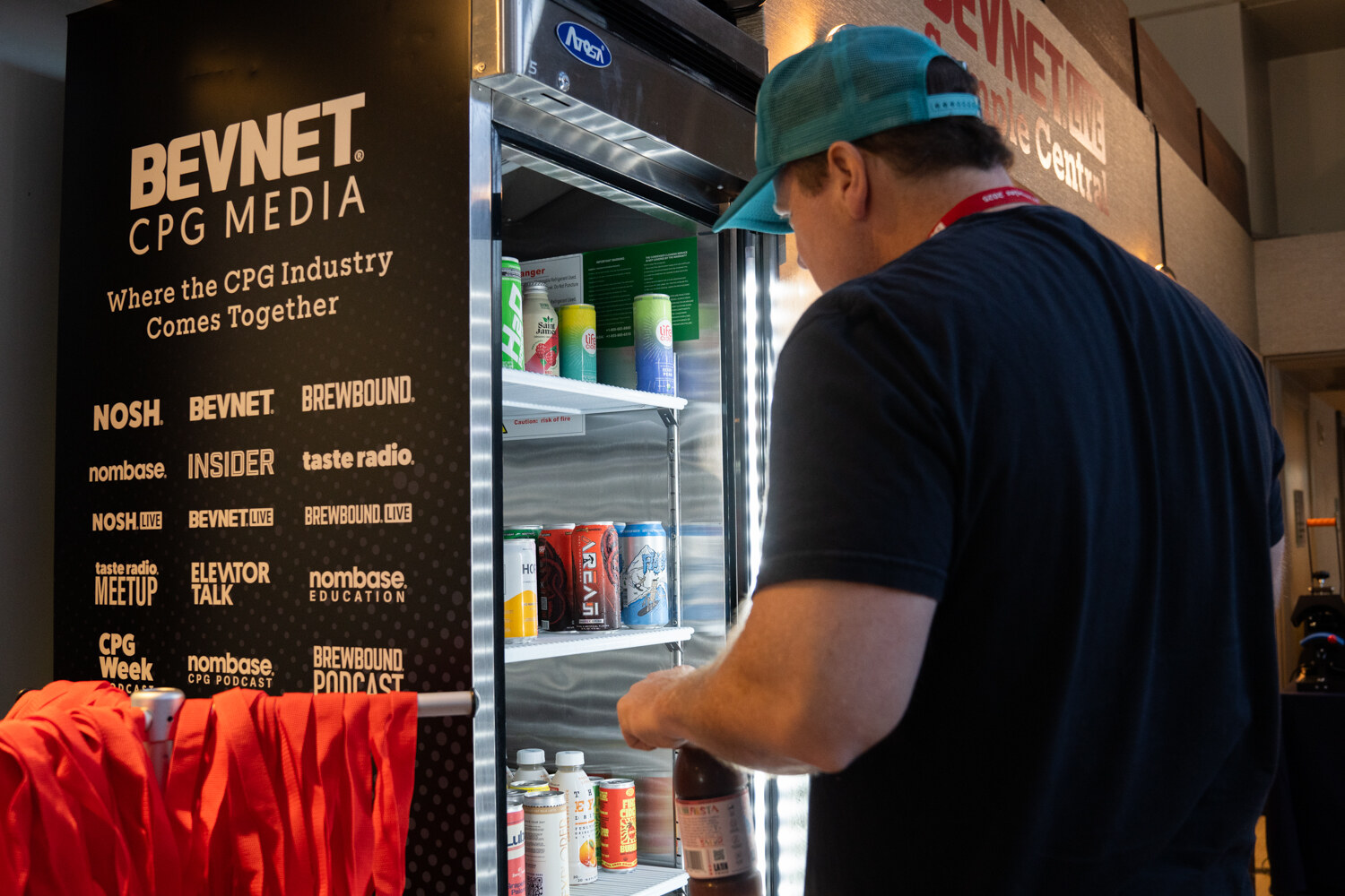 A man in a teal cap takes a drink from a BevNET CPG Media fridge.