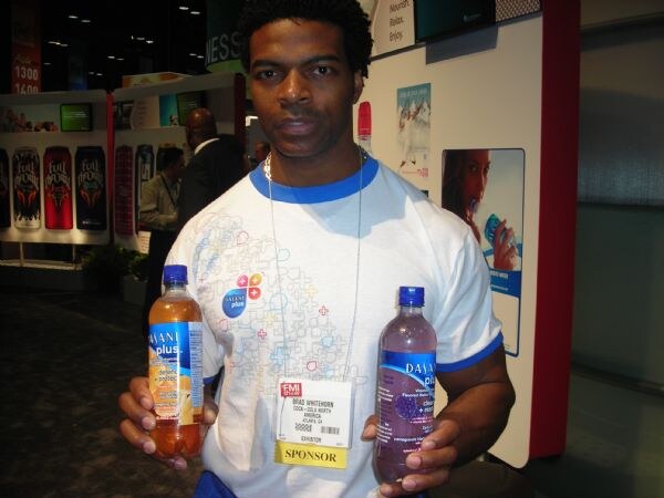 Brad Whitehorn of Coca-Cola North America, wearing a white T-shirt and a sponsor badge, holds a bottle of orange Dasani Plus in one hand and a purple Dasani Plus in the other.