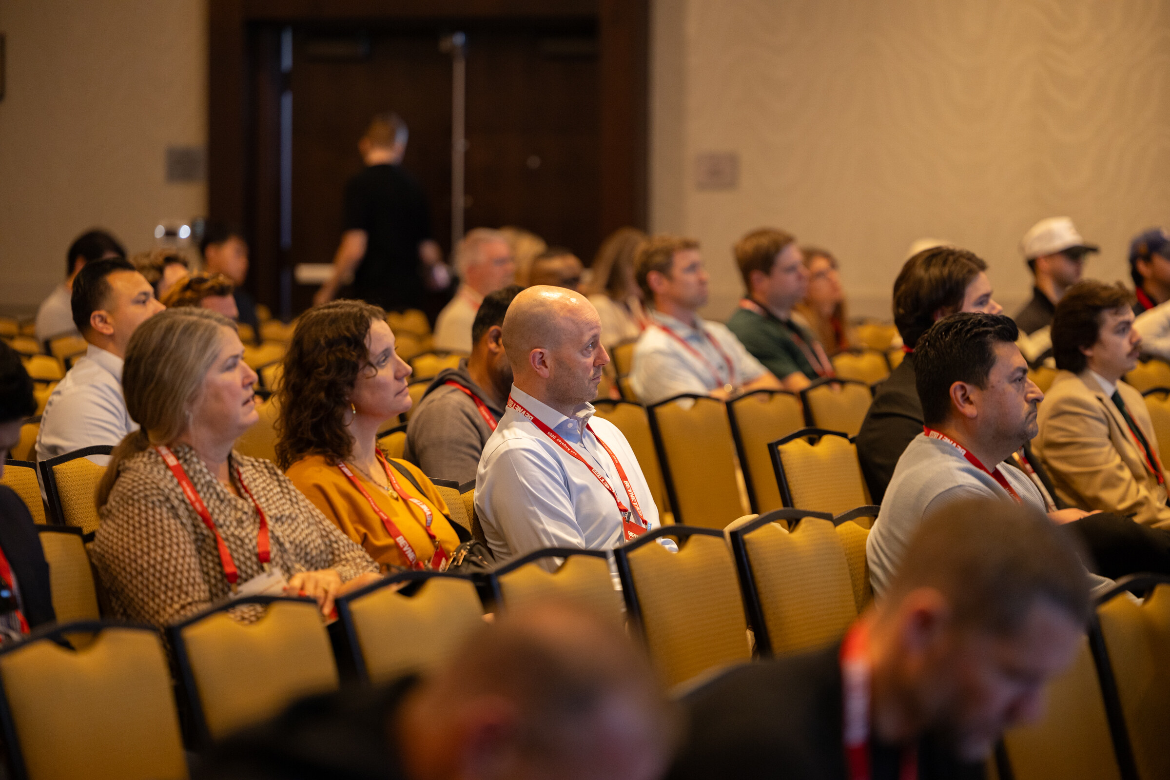 Attendees with lanyards sit in rows of gold chairs.