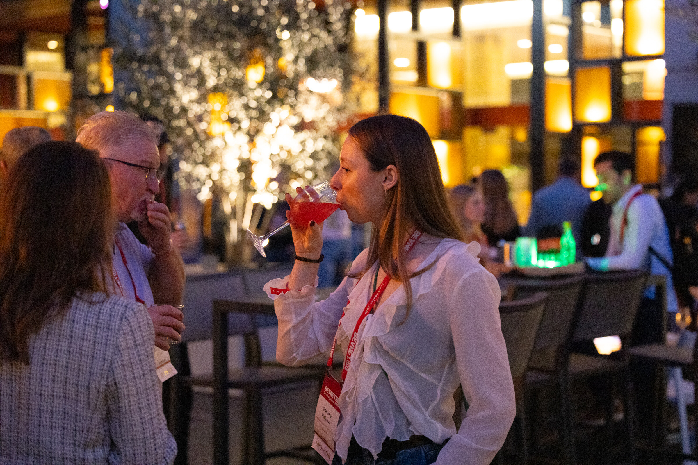 A woman in a white blouse and lanyard sips a pink drink at an evening reception.