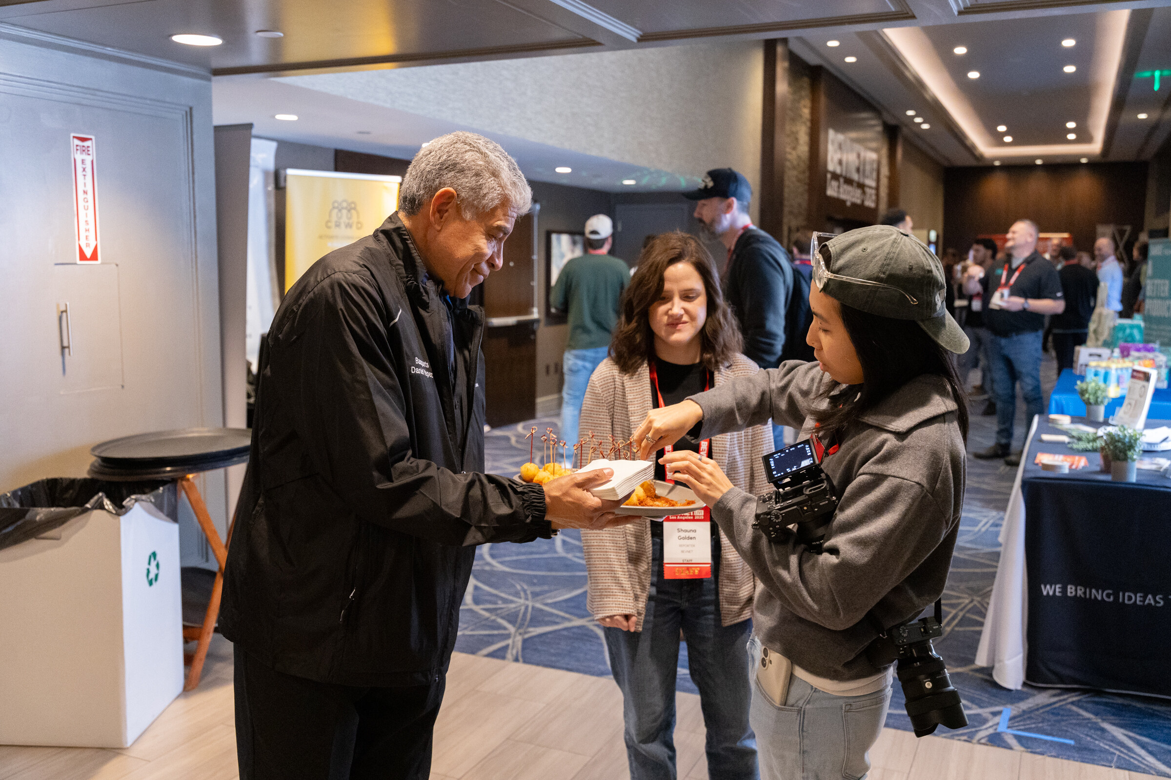 A man in a dark jacket offers hors d'oeuvres to guests while a photographer works nearby.