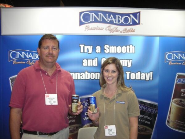 A man in a red polo and a woman in an olive Cinnabon shirt each hold a can of Cinnabon coffee latte in front of a blue Cinnabon Premium Coffee Lattes banner.