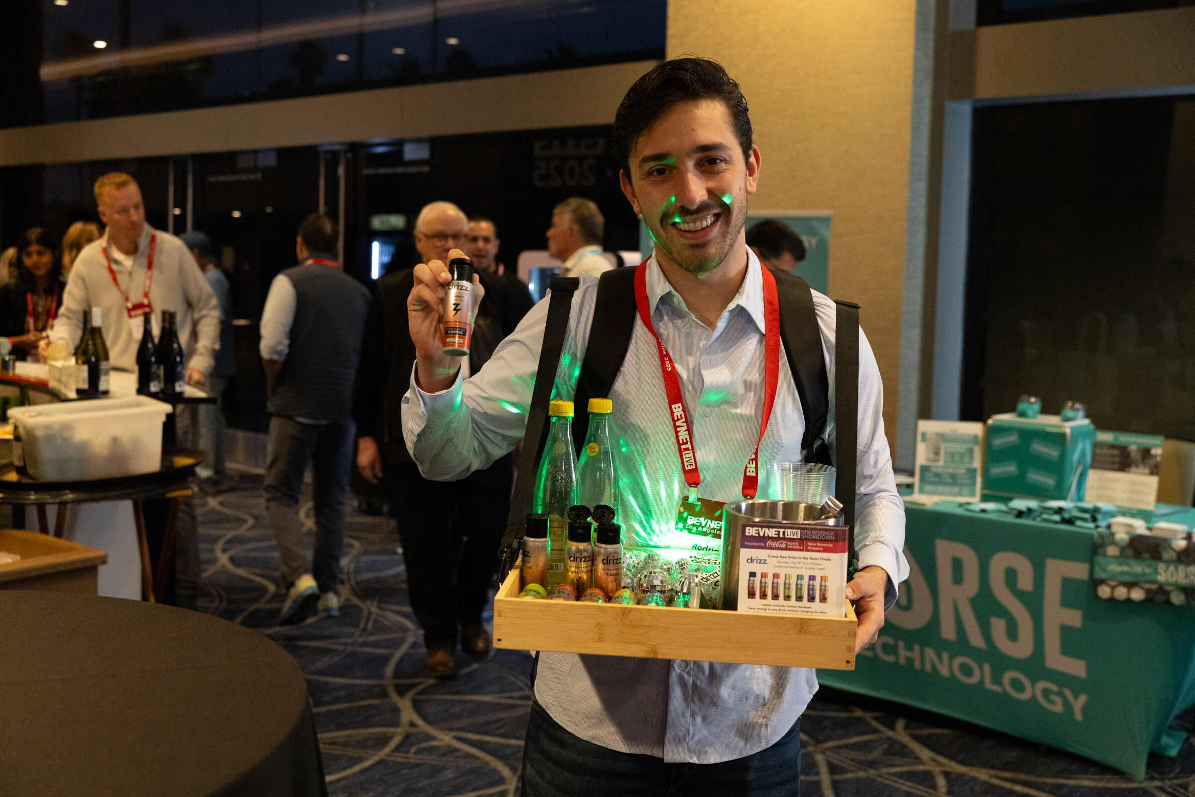 A smiling man holds a wooden tray of small glowing bottles in a busy reception room.