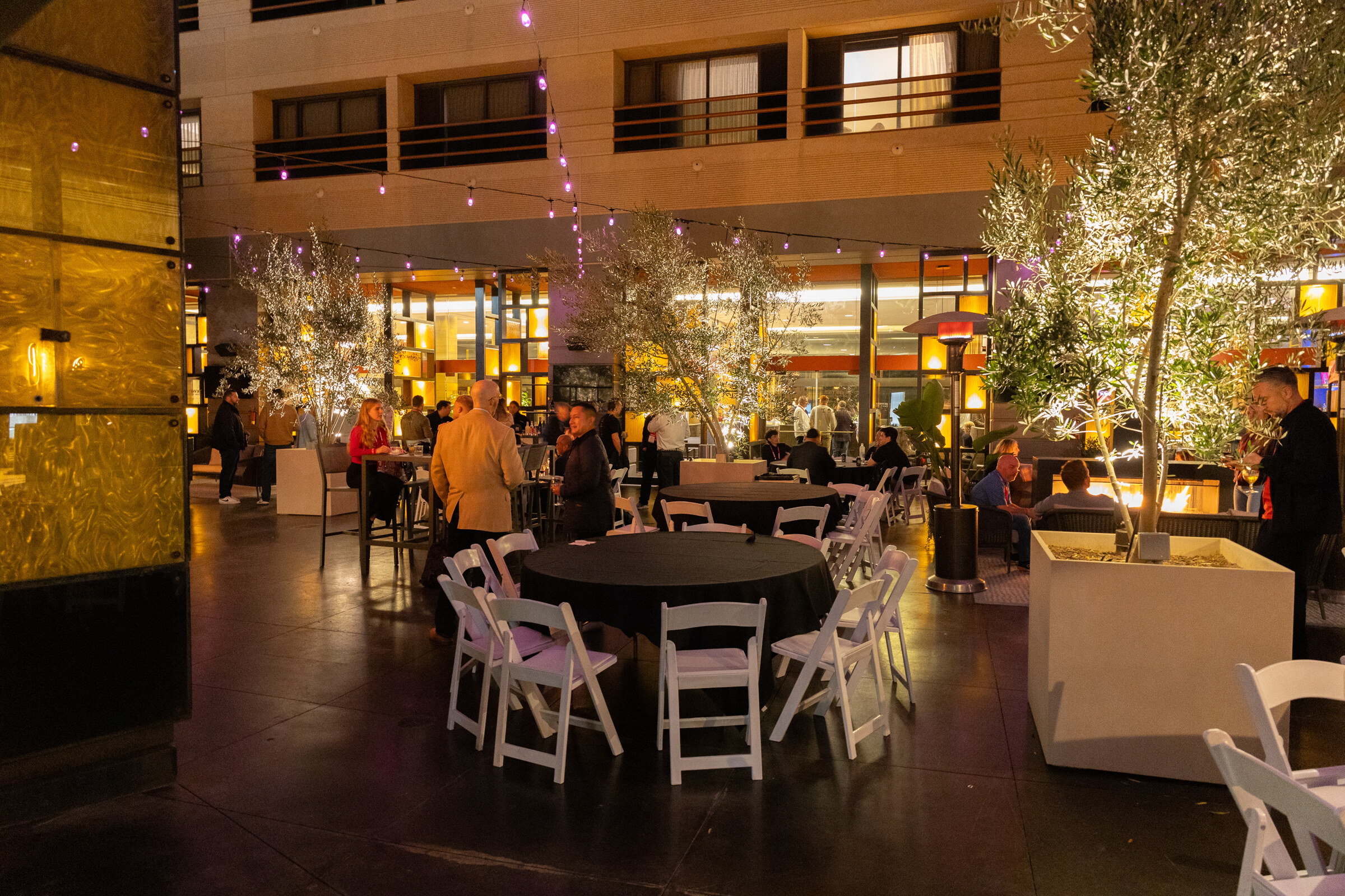 Round tables and white folding chairs sit in a lit courtyard with trees at night.