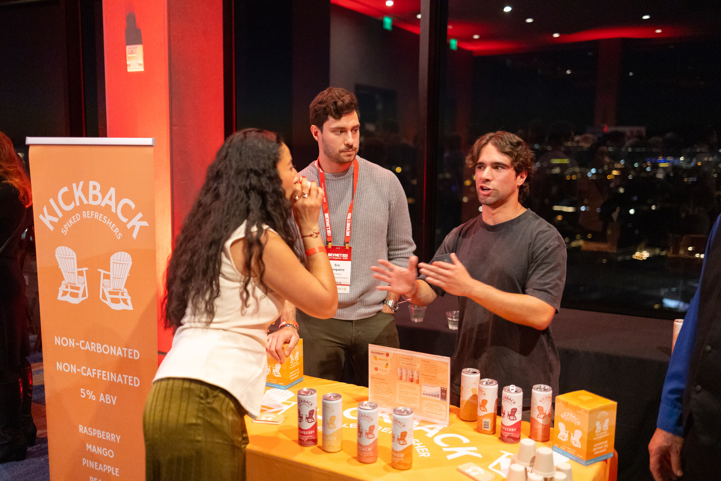A woman and two men talk at an orange Kickback booth with cans on the table.
