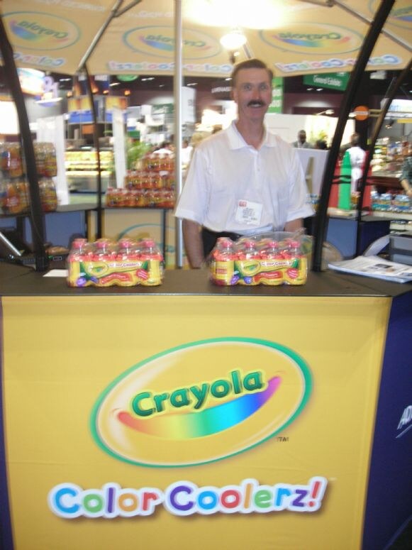 A mustached man in a white shirt stands behind a yellow counter marked Crayola Color Coolerz, with two multipacks of Color Coolerz bottles on top.