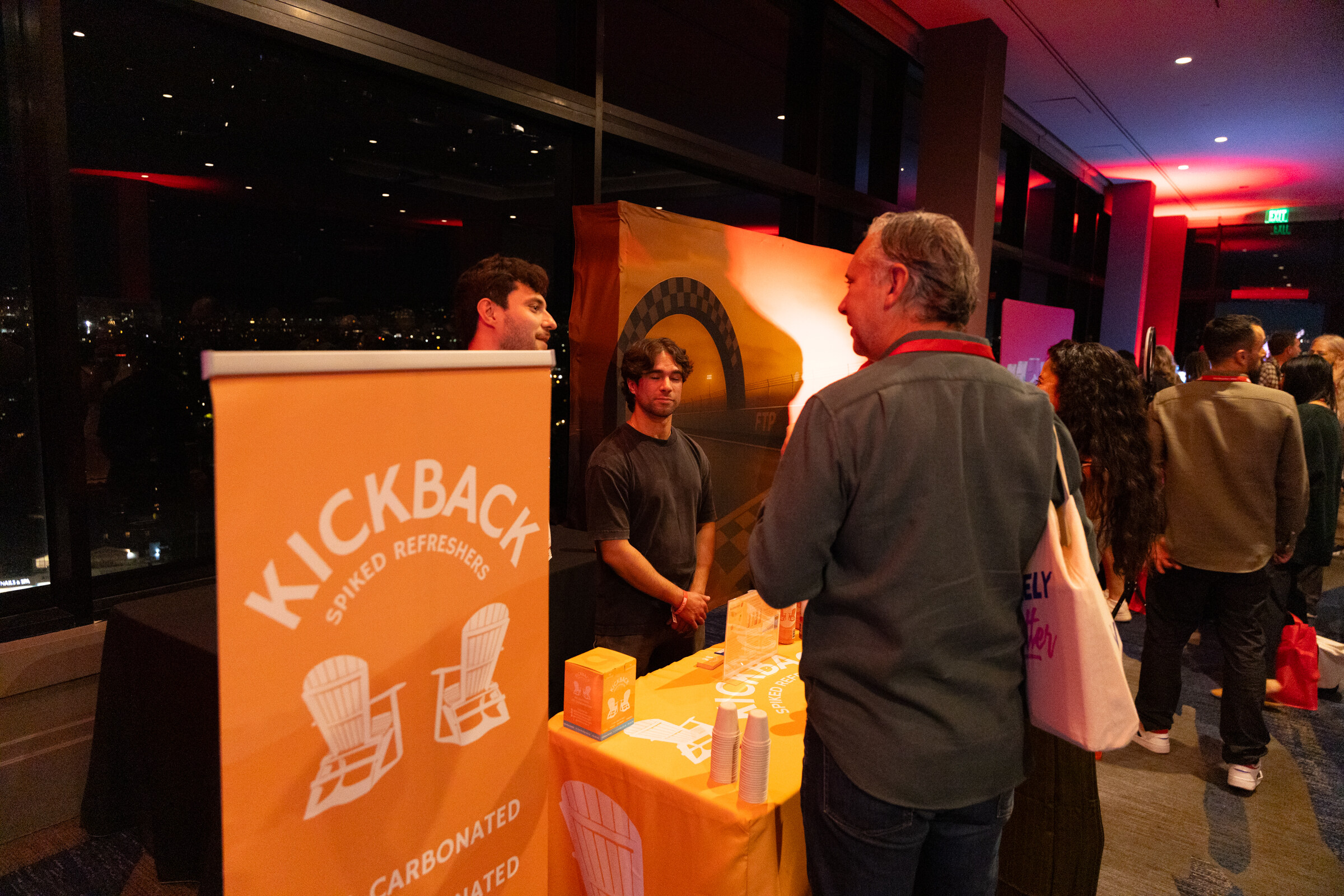 A man talks to someone at an orange Kickback booth against a night window.