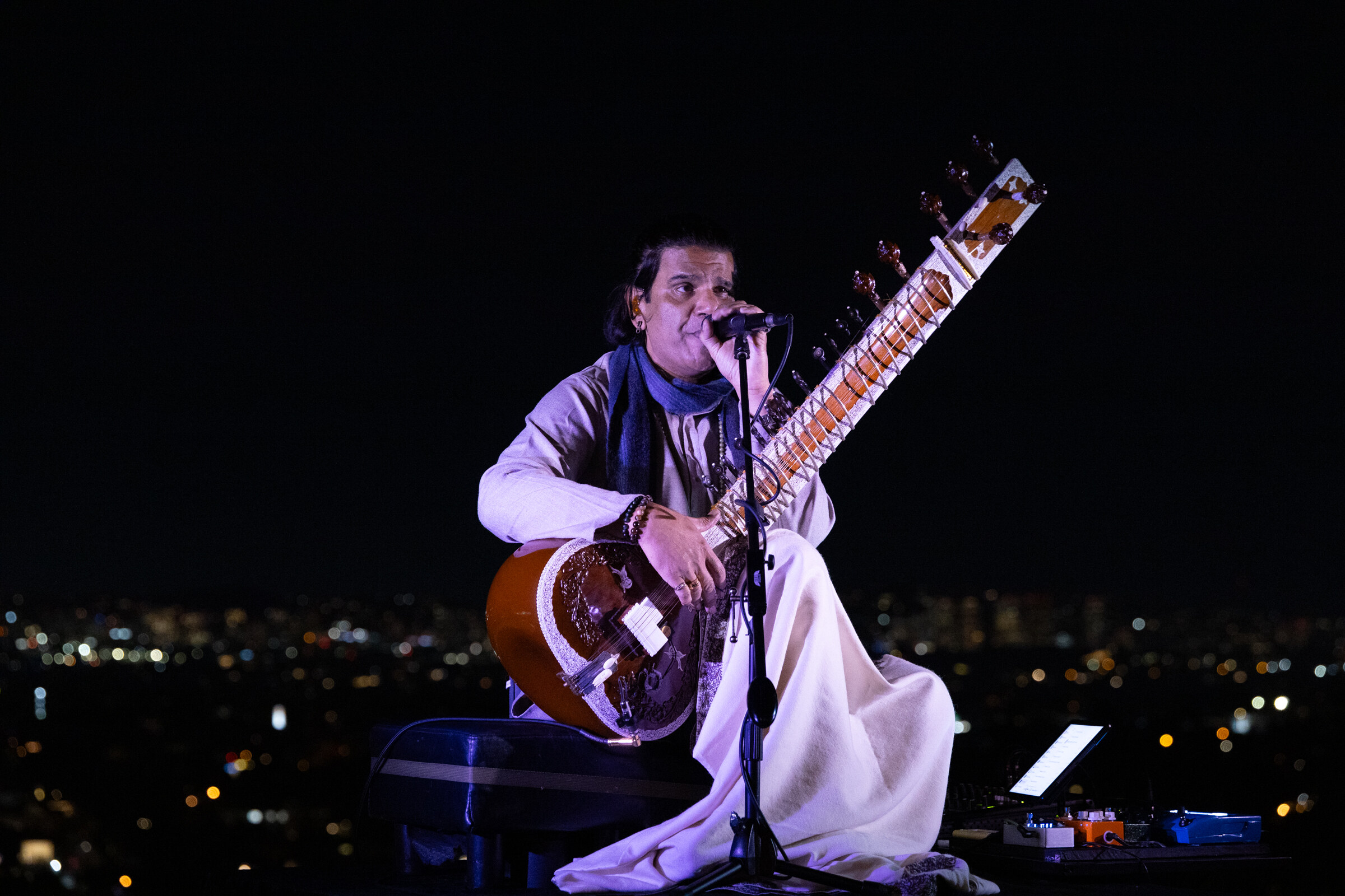 A musician in white plays a sitar and sings against a dark city skyline.