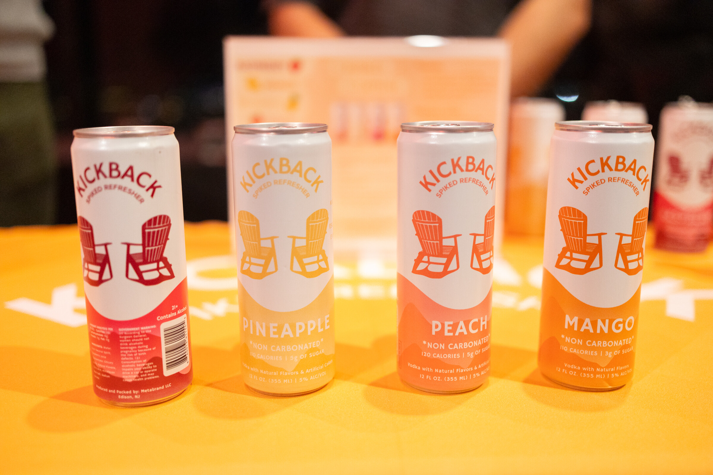 Four Kickback cans in pineapple, peach and mango flavors stand on an orange table.