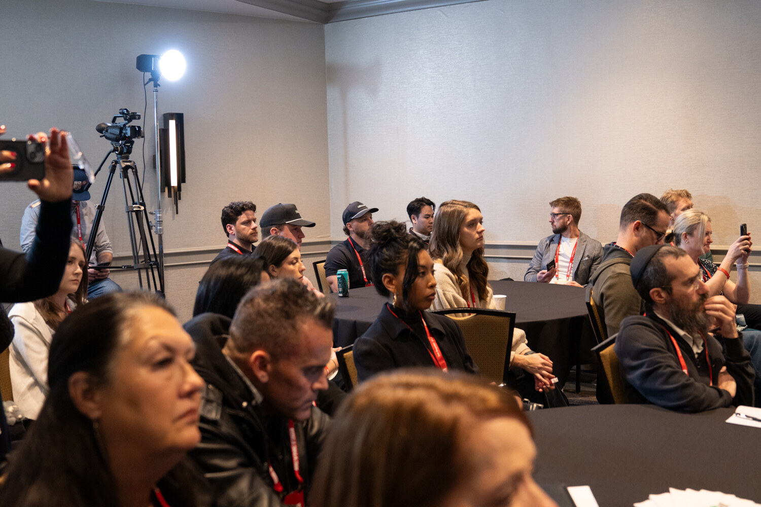 Attendees seated at tables listen to a presenter, with a video camera on a tripod behind them.