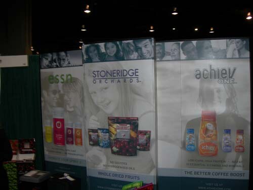 A wide view of three booth panels for essn, Stoneridge Orchards and achiev ONE, with product images and photos of people, under ceiling lights in an exhibit hall.