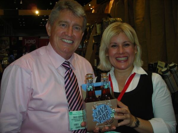 David Cooper, at left in a pink shirt and striped tie, stands beside a blonde woman who holds a four-pack of Java Pop vanilla coffee soda. Both are smiling.