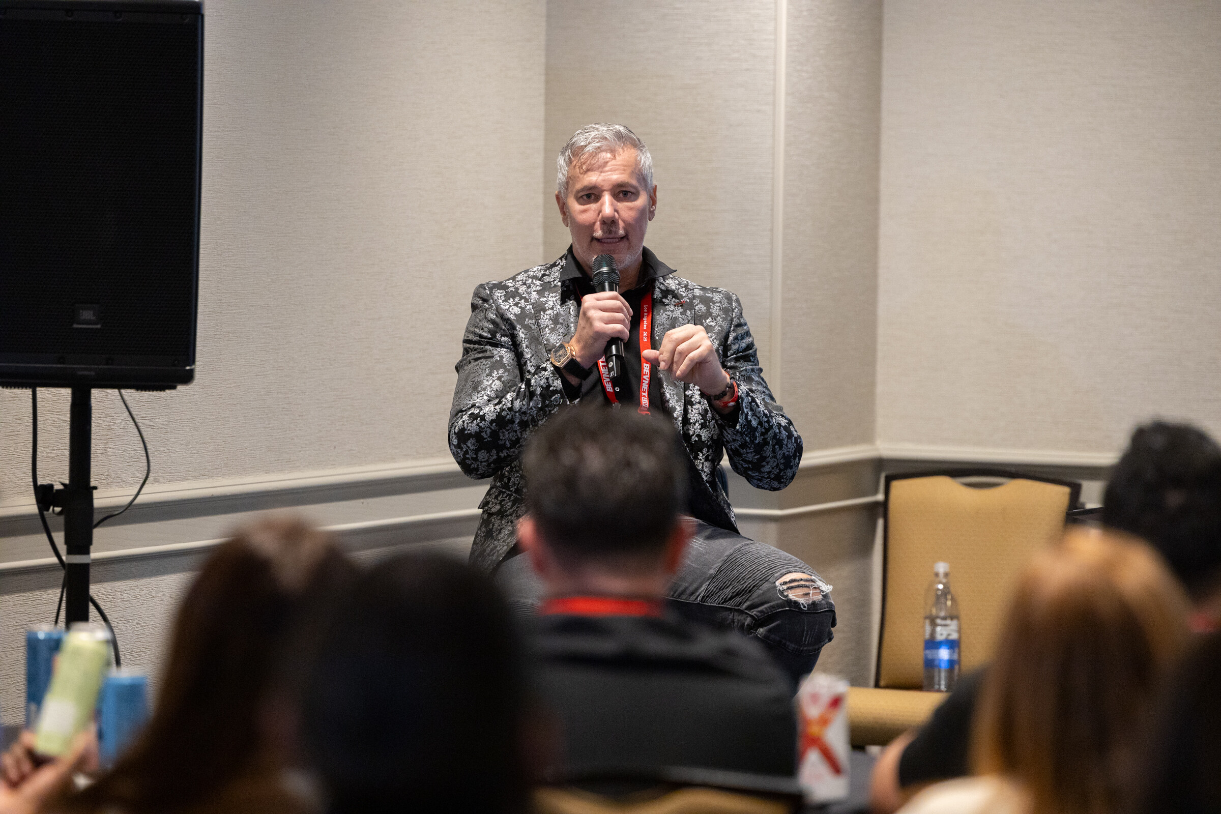 A man in a patterned jacket speaks to a seated room.