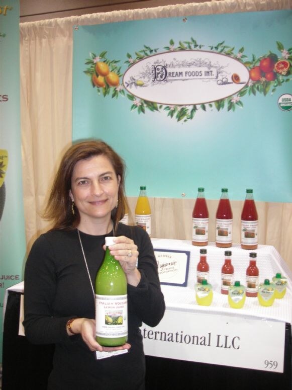 A smiling woman in a black top holds a bottle of Italian Volcano lemon juice. Behind her is a Dream Foods Int. sign and a table of juice bottles and small lemon juice containers.