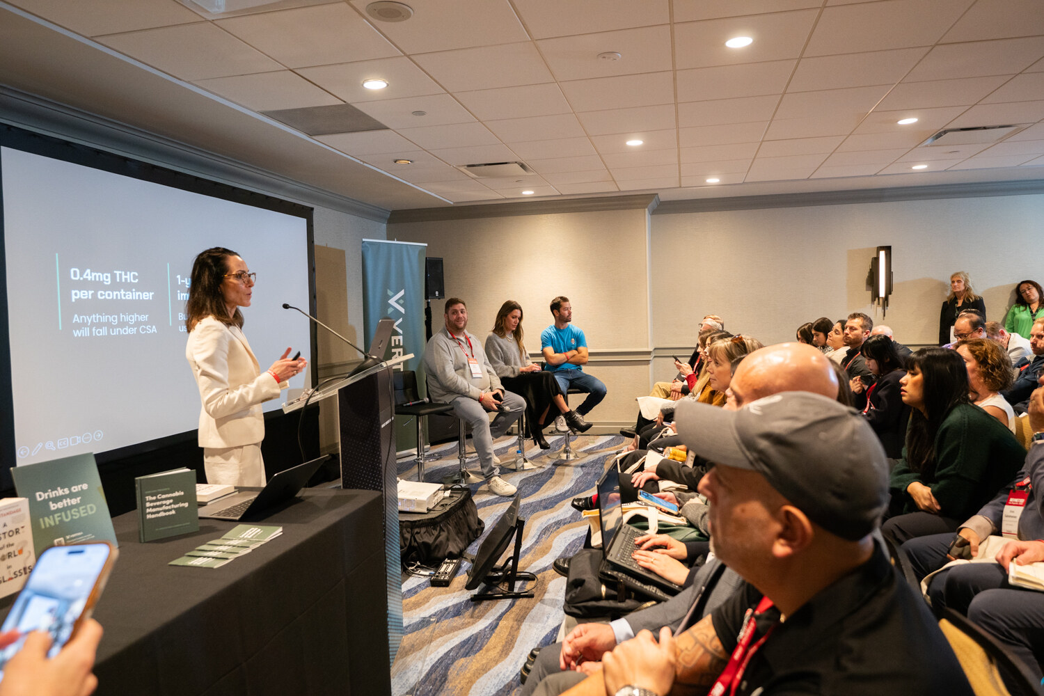 A woman presents while panelists sit and attendees watch in a meeting room.