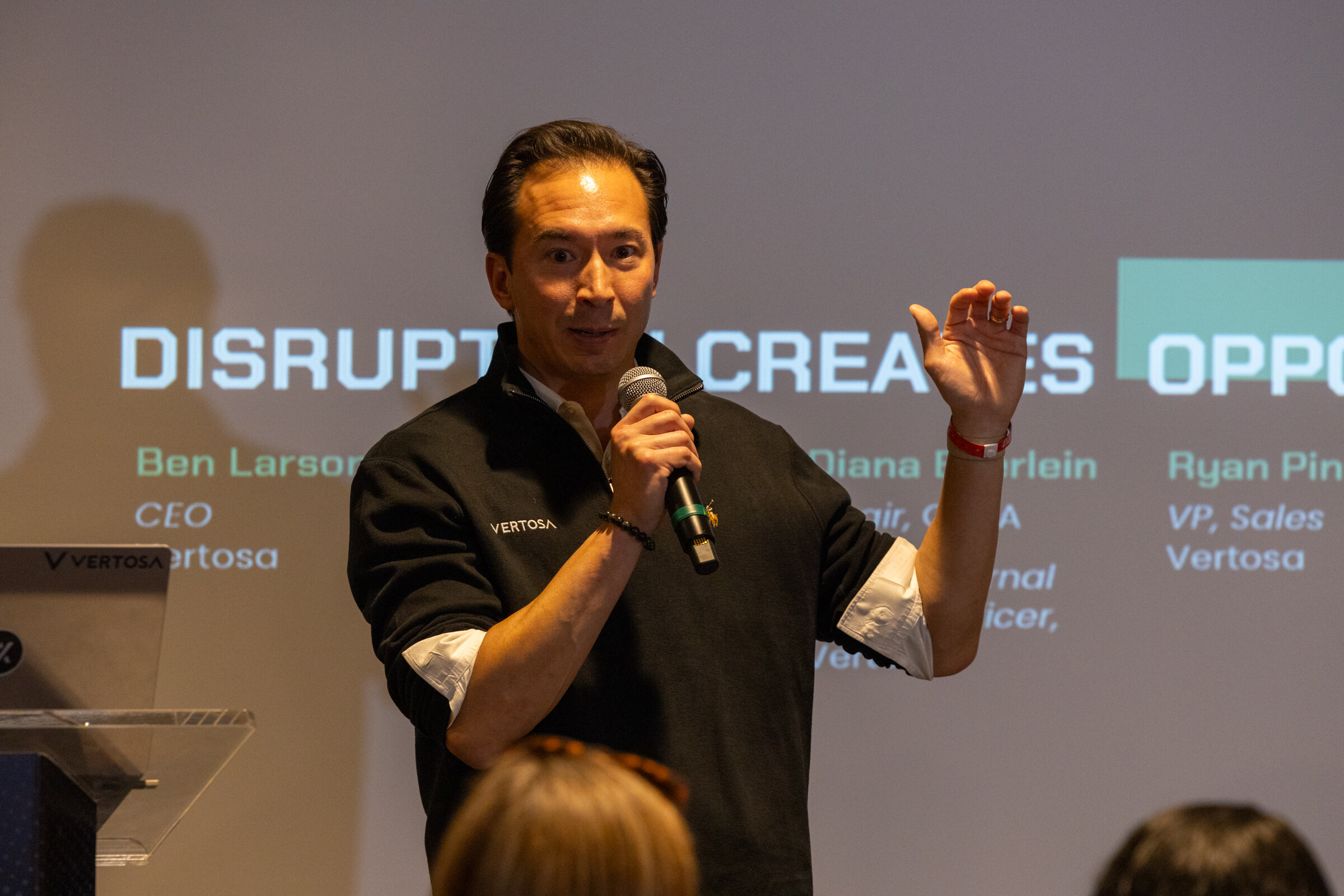 A man in a black quarter-zip speaks into a microphone in front of a slide.