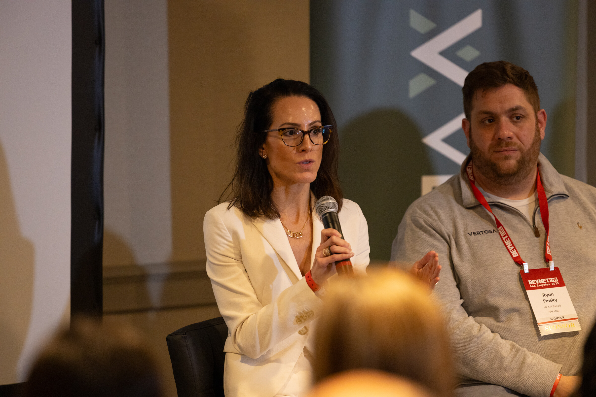 A woman in a white blazer and a man in a gray sweater speak into microphones.