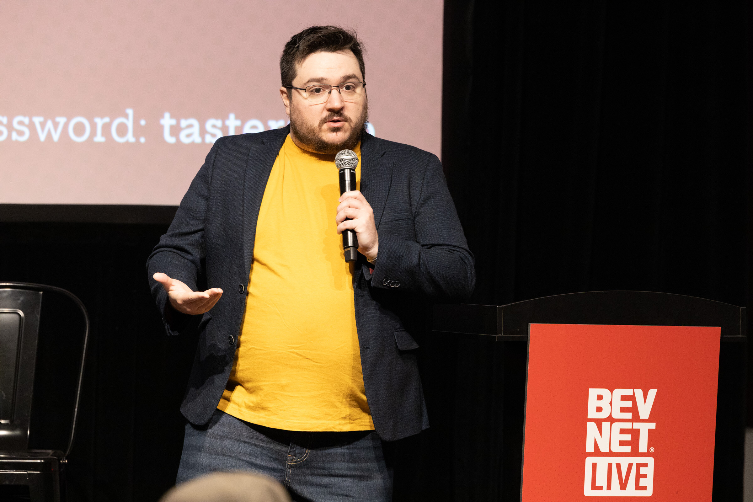 Man in a navy blazer and yellow T-shirt speaking into a microphone beside a red BevNET Live podium.