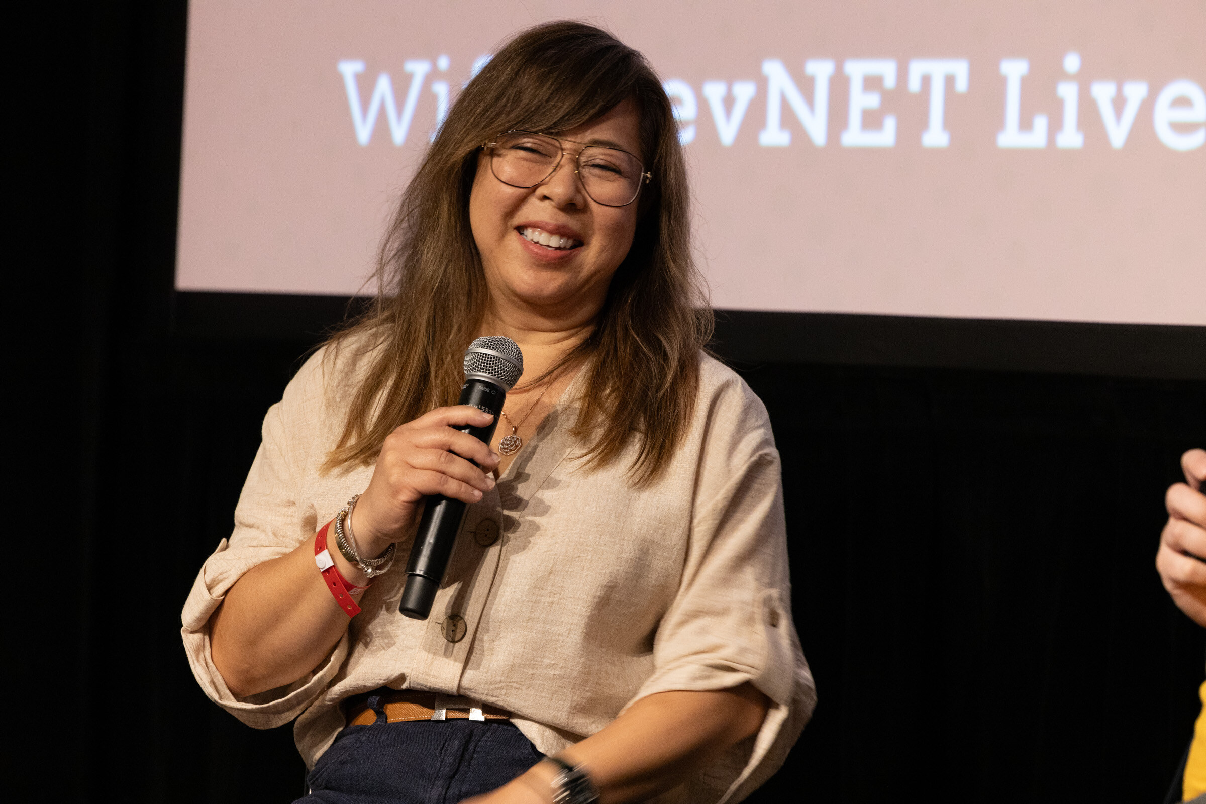 Smiling woman in glasses and a linen shirt holding a microphone on stage.