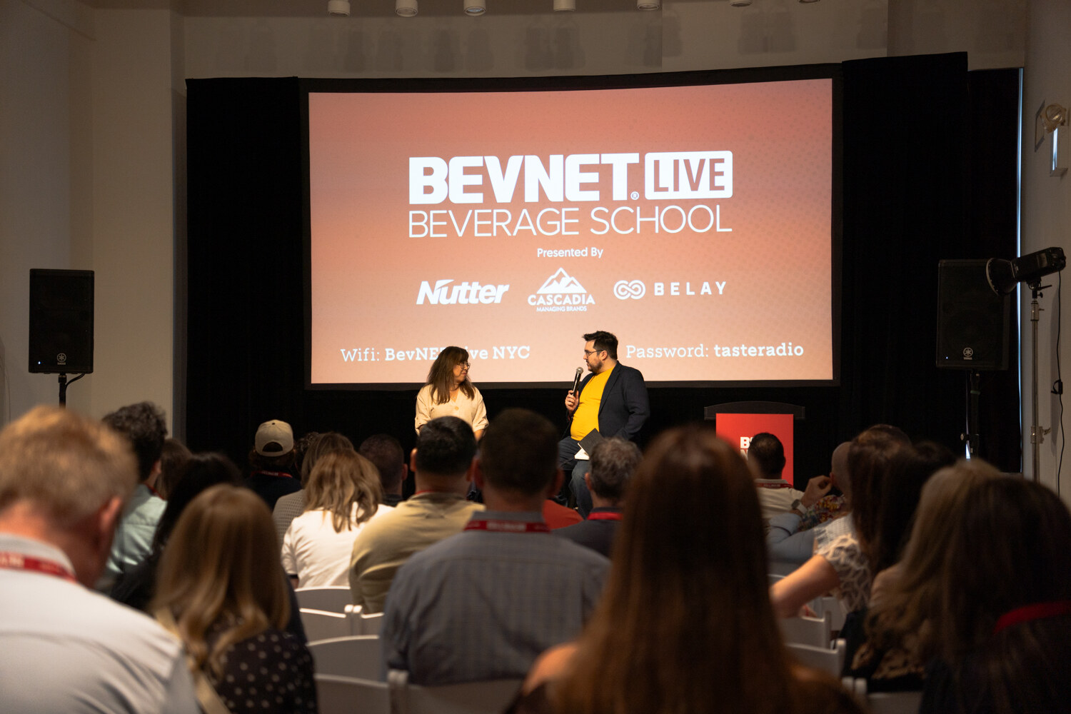 Audience facing a stage with two people seated before a large BevNET Live Beverage School slide.