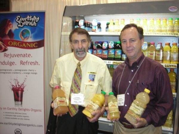 Two men holding bottles of juice stand beside an Earthly Delights Organic banner and a refrigerated case of bottled juices.