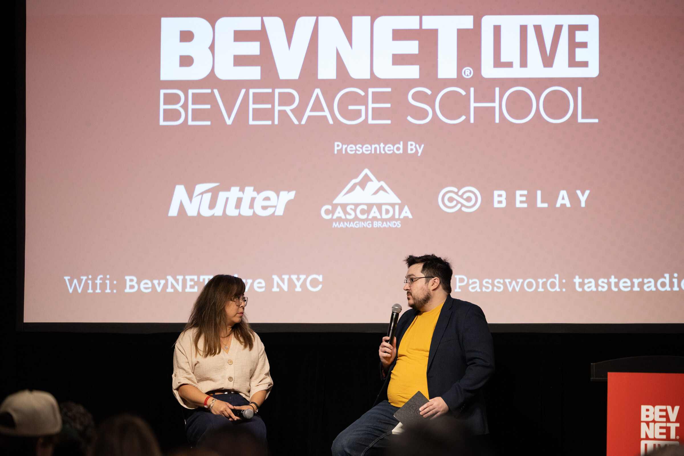 Two people seated on stage in conversation beneath a slide reading “BevNET Live Beverage School, presented by Nutter, Cascadia, Belay.”