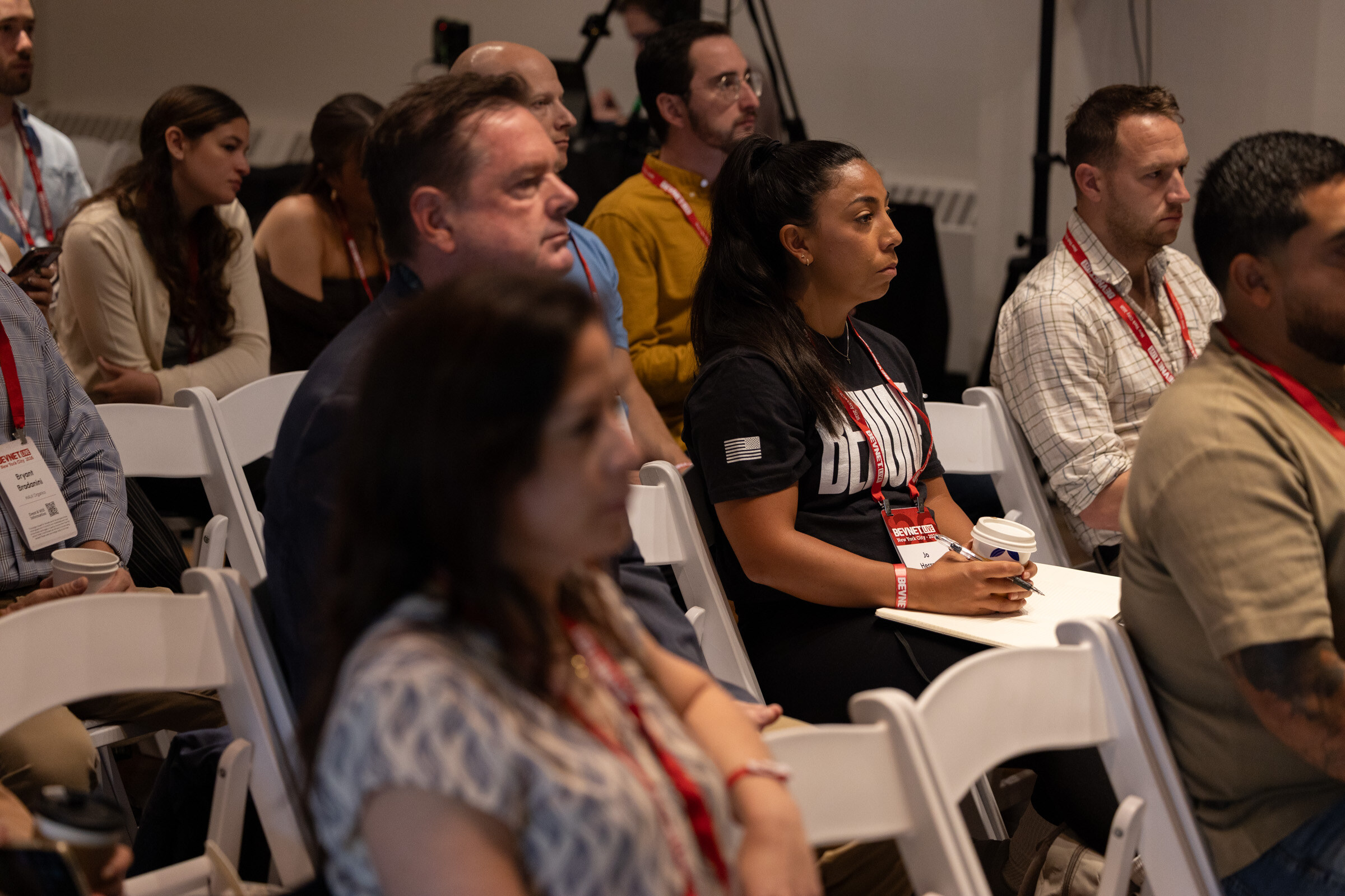 Attendees with lanyards seated in white folding chairs, several taking notes.